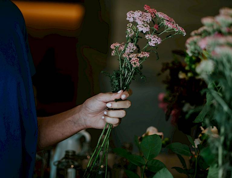 Una persona tiene in mano un mazzo di fiori rosa, circondato da altri arrangiamenti floreali.
