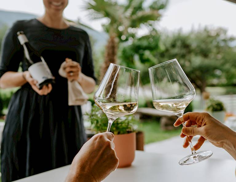 Due persone brindano con bicchieri di vino bianco mentre una donna serve una bottiglia all'aperto.
