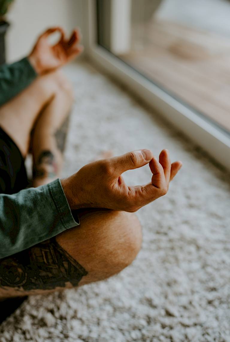 Persona seduta a gambe incrociate che medita davanti a una finestra su un tappeto, con le mani in Mudra.