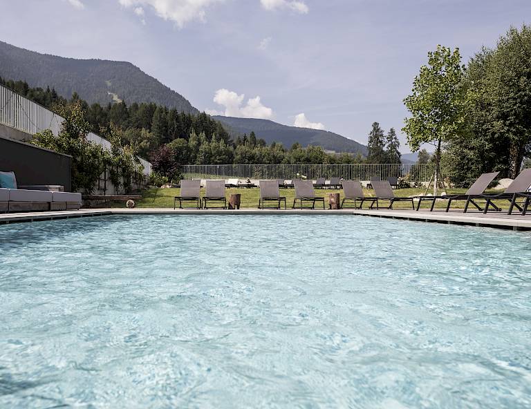 Piscina all'aperto con lettini, circondata da montagne e alberi sotto un cielo azzurro.