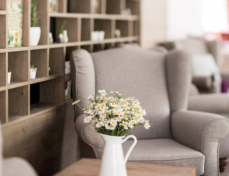 Sala lettura accogliente con poltrona grigia, libreria di legno e fiori bianchi in vaso.