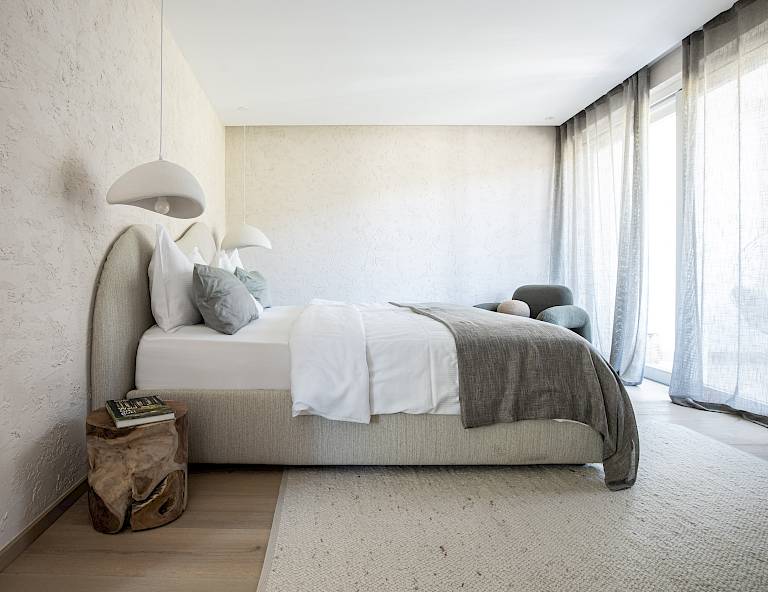 Camera da letto moderna con letto grande, lampada sospesa, pavimento in legno e finestre ampie che lasciano entrare molta luce.