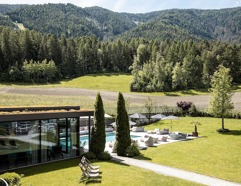 Villa moderna con facciata in vetro, piscina e giardino, circondata da paesaggio verde e bosco.