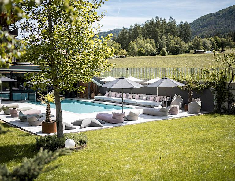 Zona piscina moderna con lettini su prato verde, circondata da natura e alberi.