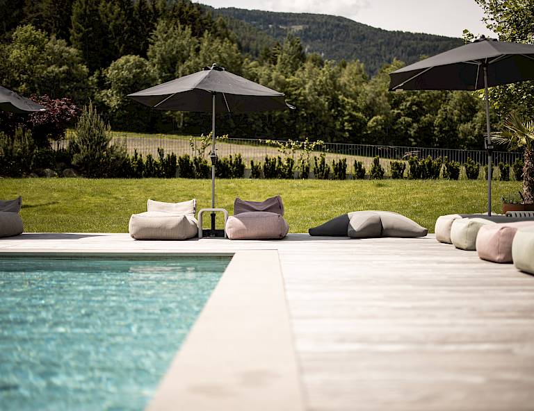 Area piscina con cuscini e ombrelloni davanti a un paesaggio verde e colline boscose sullo sfondo.