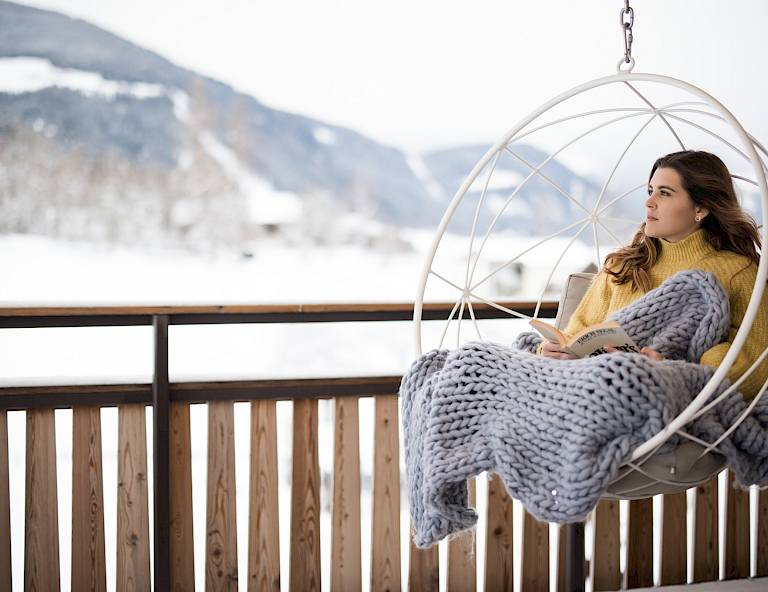 Donna seduta su una sedia sospesa su un balcone, avvolta in una coperta, con montagne innevate sullo sfondo.