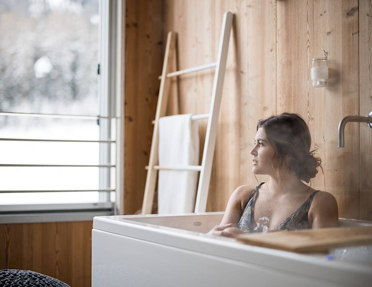 Donna rilassata nella vasca da bagno, guarda fuori dalla finestra. Pareti di legno e asciugamani sullo sfondo.