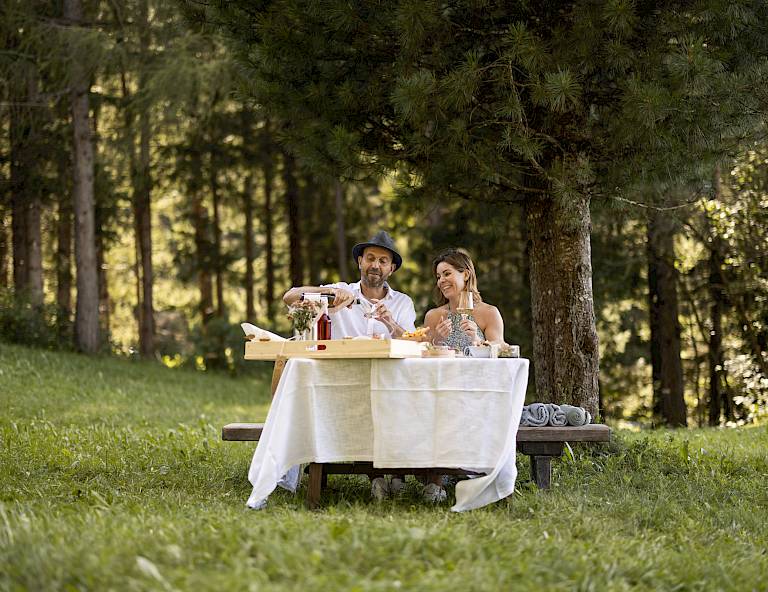 Coppia che si gode un picnic nel bosco, seduta a un tavolo sotto alti alberi. Raggi di sole tra le foglie.