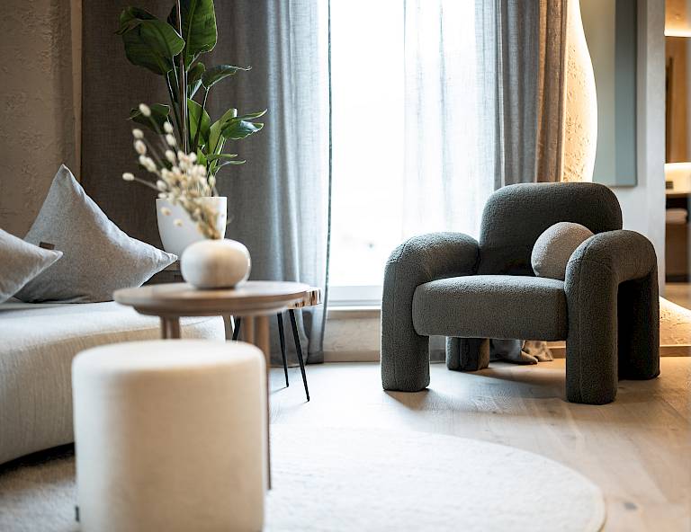 Soggiorno moderno con poltrona grigia, tavolino rotondo, cuscini decorativi e piante sullo sfondo.