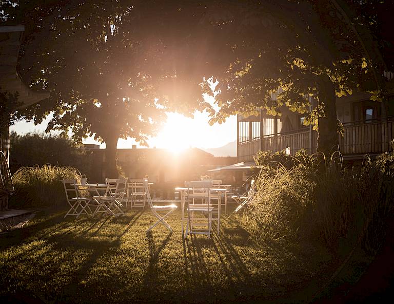 Giardino al tramonto con mobili bianchi all'ombra di un grande albero, illuminato dal sole.