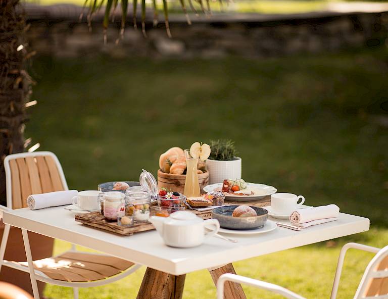 Tavola apparecchiata all'aperto con panini, marmellata e stoviglie sotto una palma in giardino.