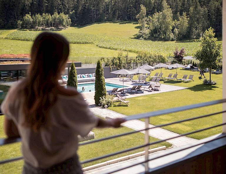 Una donna su un balcone guarda un giardino con piscina e lettini, circondato da boschi.