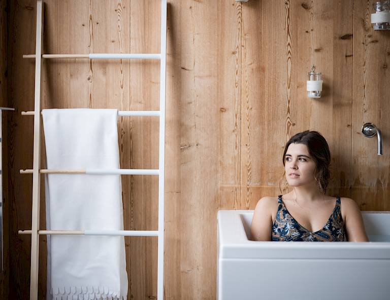 Donna si rilassa nella vasca da bagno, circondata da pareti in legno, con asciugamano sul portasalviette.