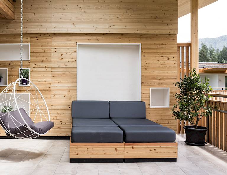 Terrazza moderna con pareti in legno, divano grigio, sedia sospesa e pianta in vaso. Boschi sullo sfondo.