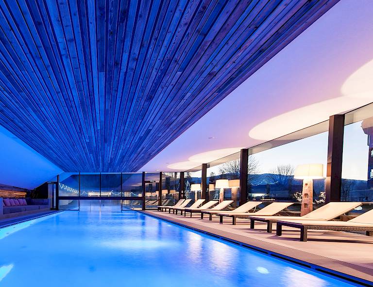 Piscina coperta di lusso con lettini moderni sotto un soffitto in legno, illuminazione blu.