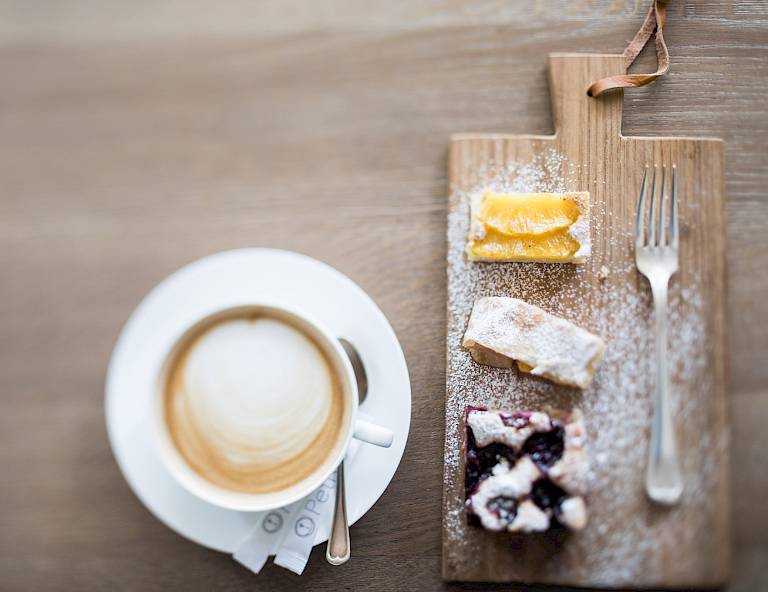 Tazza di caffè accanto a un tagliere con tre fette di torta diverse, spolverate con zucchero a velo.