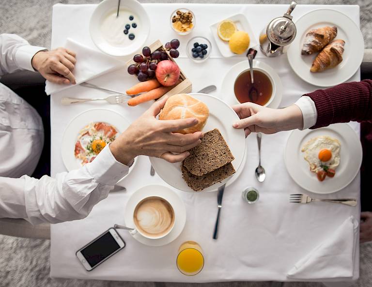 Due persone gustano una ricca colazione con pane, frutta e dolci su un tavolo bianco.
