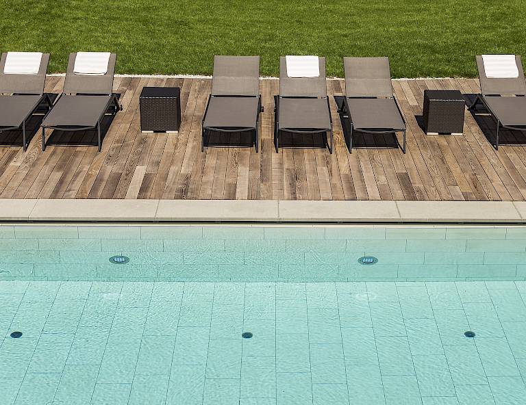Lettini a bordo piscina limpida, su una terrazza di legno con prato verde sullo sfondo.