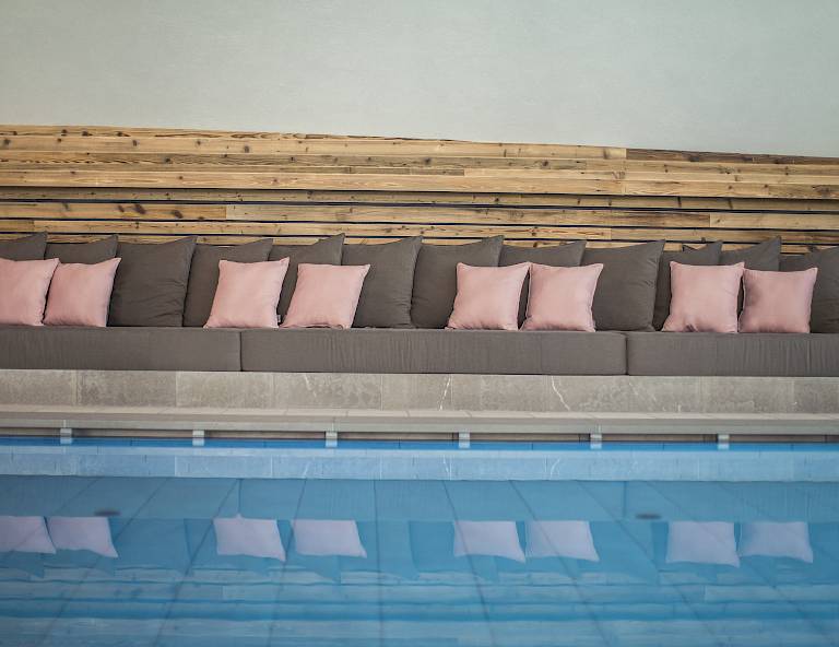 Panchina grigia imbottita con cuscini rosa riflessa sulla superficie dell'acqua di una piscina.