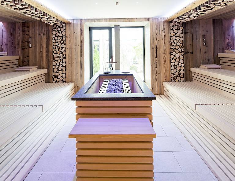 Una sauna moderna con panche di legno, rivestimenti di legno, forno centrale e cataste di legno alle pareti.