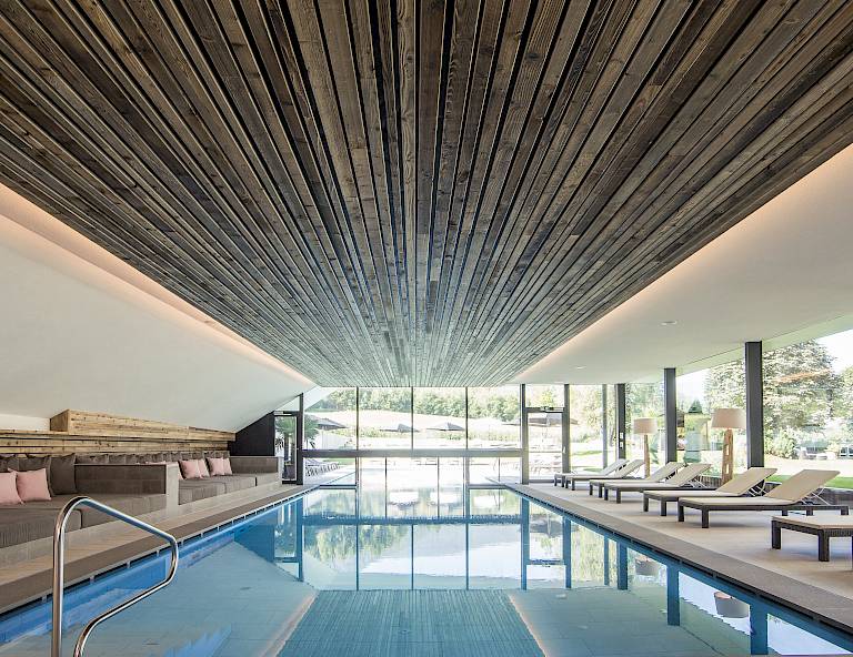 Piscina coperta moderna con soffitto in legno, lettini e grandi finestre con vista sul paesaggio.