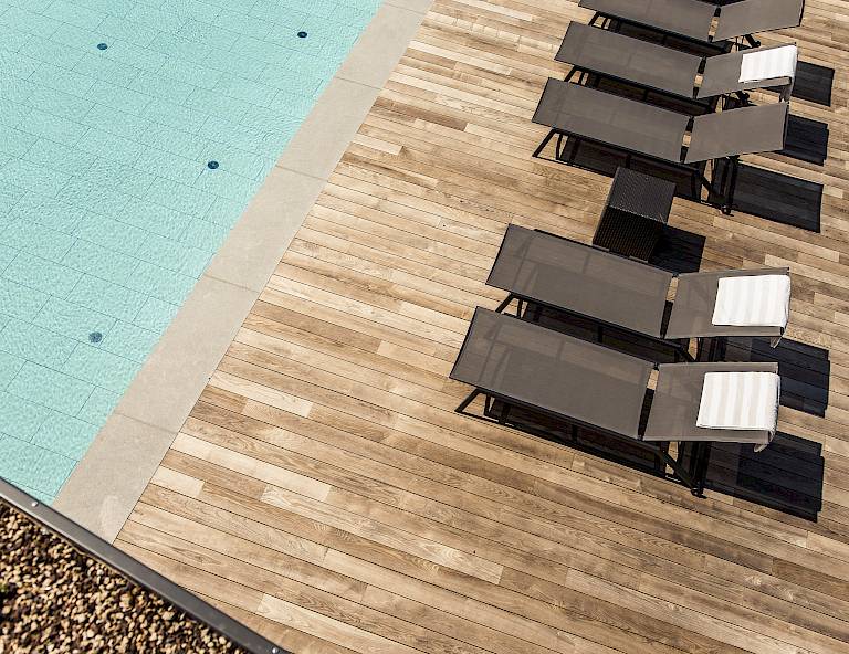 Piscina con terrazza in legno, diverse sdraio con asciugamani, luce solare intensa.