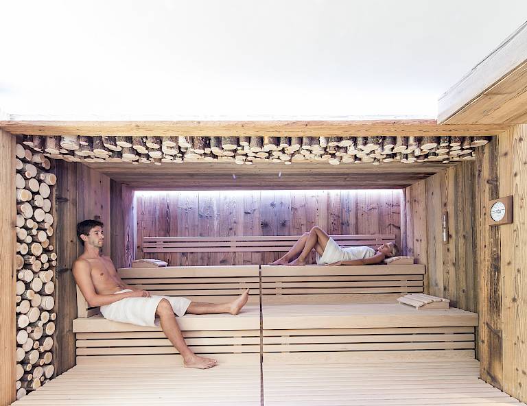 Due persone si rilassano su panche di legno in una sauna moderna, circondate da legna accatastata.