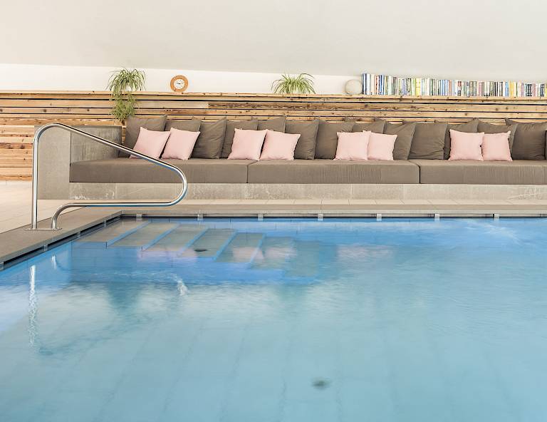 Piscina coperta moderna con comodi divani e cuscini rosa sullo sfondo. Parete in legno e libreria sopra.