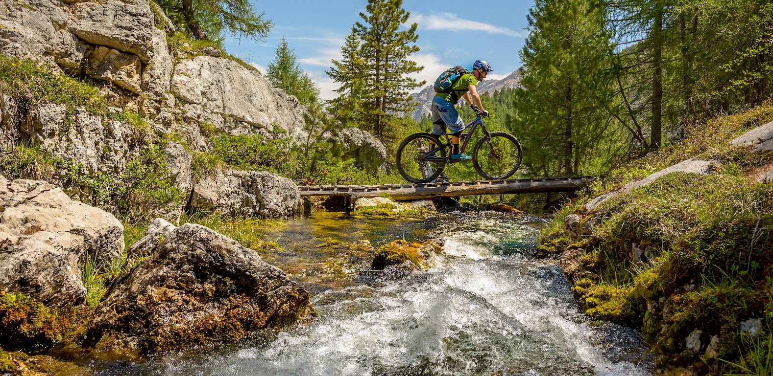 Radfahrer überquert eine schmale Holzbrücke über einen Bach in einer malerischen Berglandschaft.