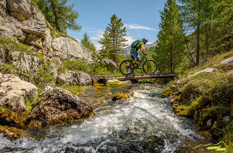 Radfahrer überquert eine schmale Brücke in malerischer Berglandschaft zwischen Bäumen und fließendem Wasser.