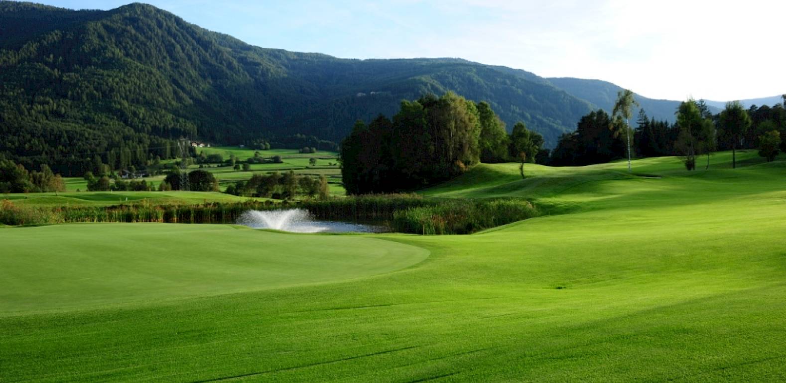 Campo da golf con prato curato, stagno e fontana, circondato da montagne sotto un cielo azzurro.