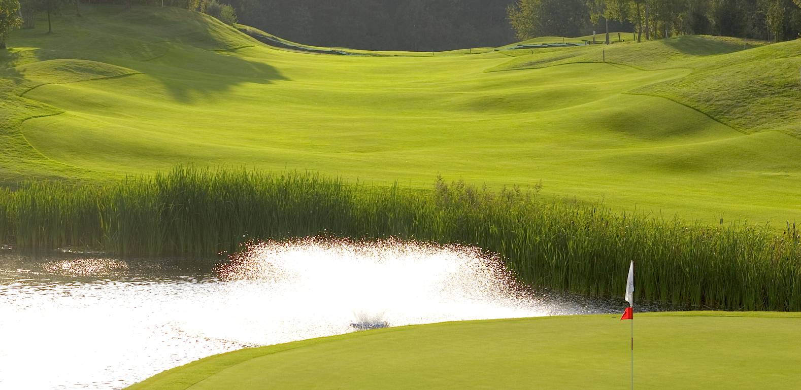 Campo da golf ben curato con uno stagno in primo piano e una bandiera bianca sul green.
