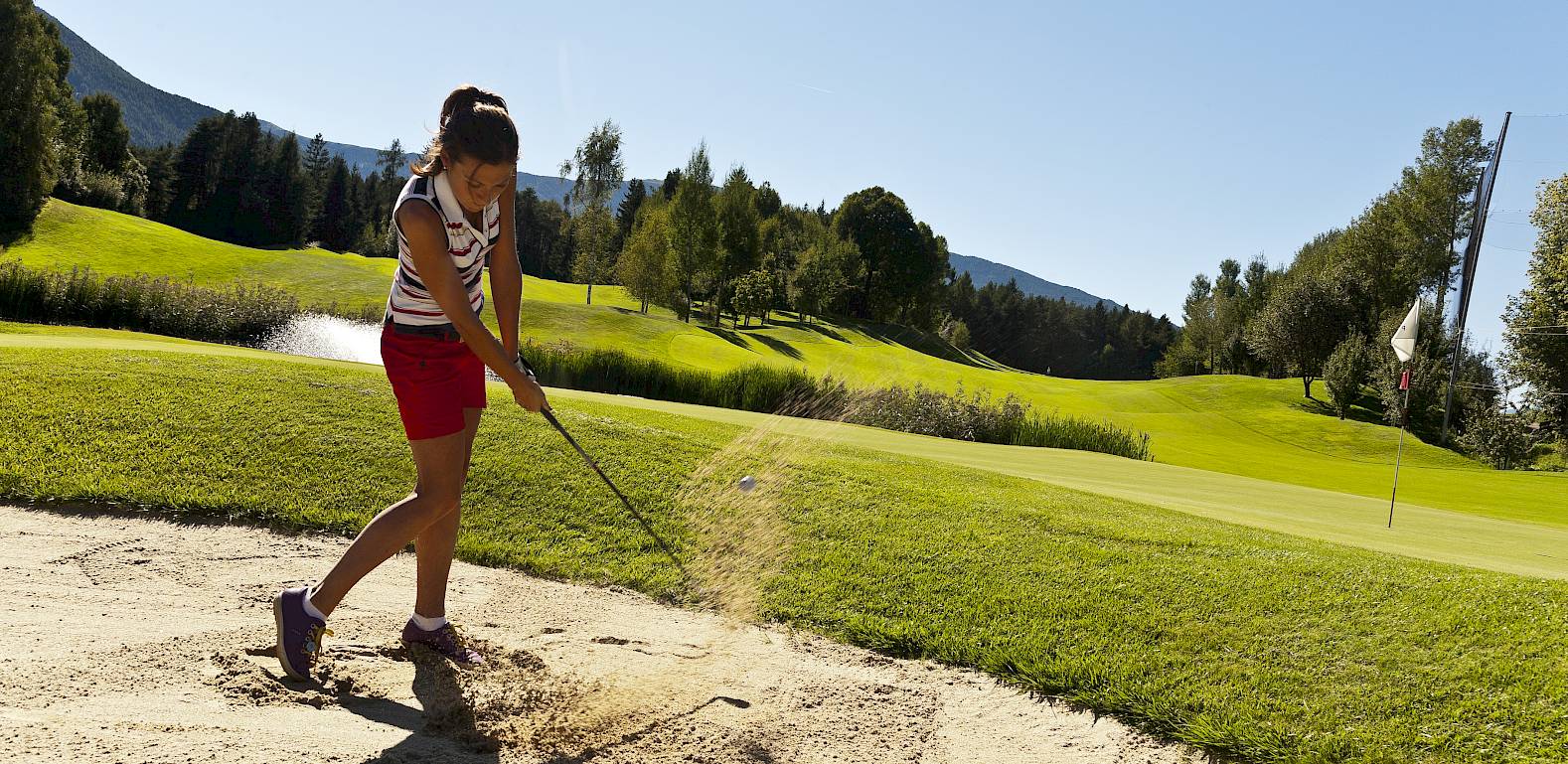 Una ragazza colpisce una pallina da un bunker su un campo da golf soleggiato con colline verdi.