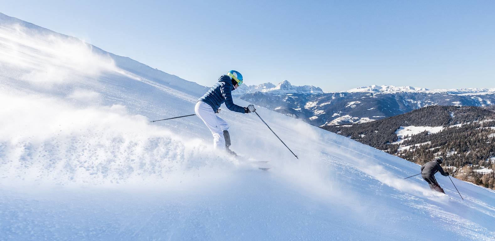 Skifahrer gleiten dynamisch die verschneite Piste hinunter, mit einer malerischen Berglandschaft im Hintergrund.