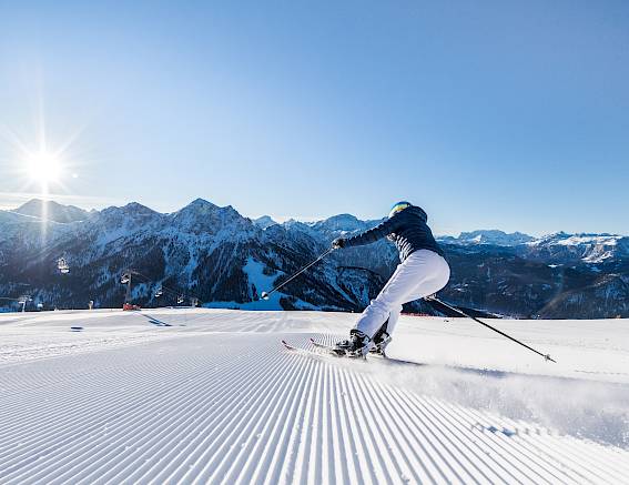 Skier on freshly groomed slope in sunshine, surrounded by snow-covered mountains.