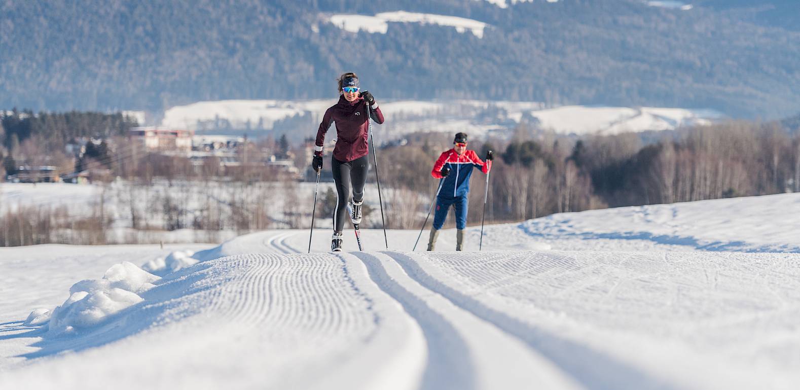 Due persone che fanno sci di fondo su pista preparata; montagne innevate sullo sfondo.