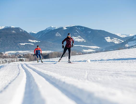 Two cross-country skiers on groomed snow track, surrounded by mountains and blue sky.