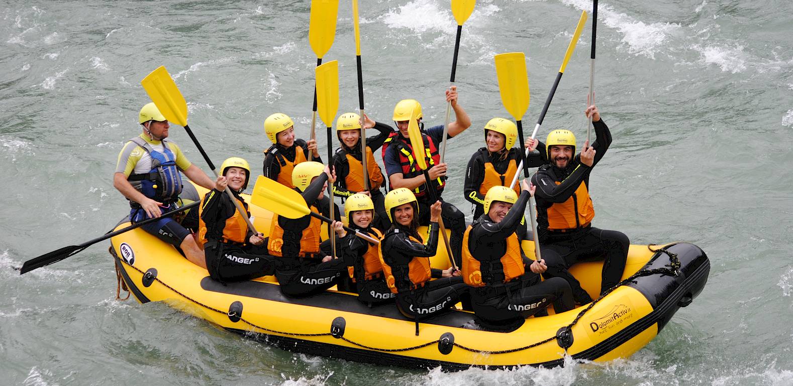 Menschen in gelben Helmen beim Rafting in einem Fluss, heben Paddel in die Luft und freuen sich.