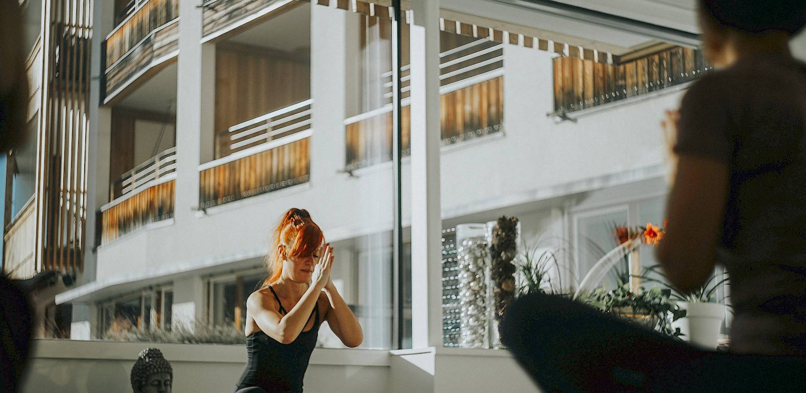 Una persona fa yoga in una stanza moderna con luce intensa e grandi finestre. Si vede una statua di Buddha.