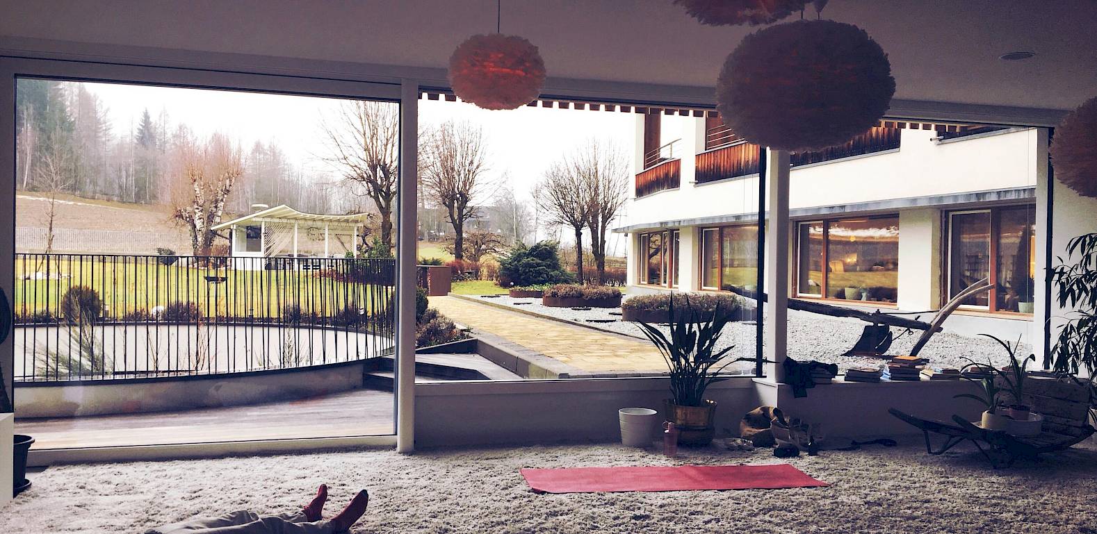 Interno con vista esterna, persone distese su tappeto, grandi finestre mostrano giardino e alberi.