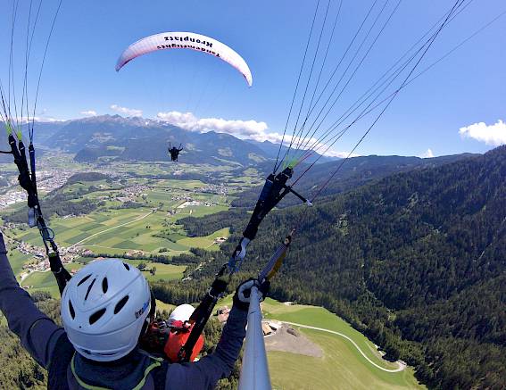 Paragleiter fliegen über eine grüne Landschaft mit Bergen im Hintergrund, blauer Himmel und Wolken.