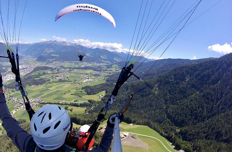 Paragleiter fliegen über eine grüne Landschaft mit Bergen im Hintergrund, blauer Himmel und Wolken.