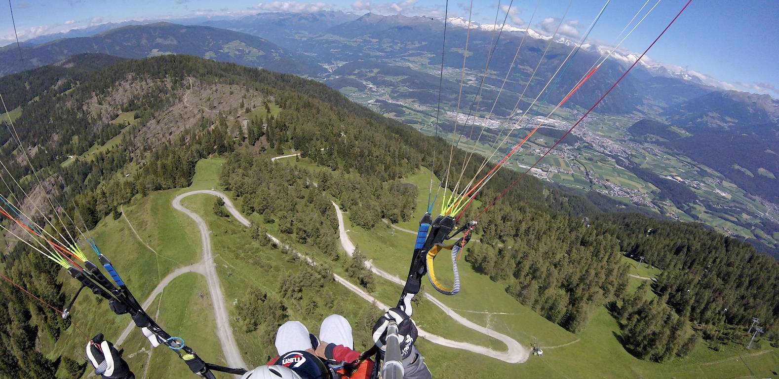 Parapendio sopra paesaggio montano verde con strade tortuose e vista sulla valle.