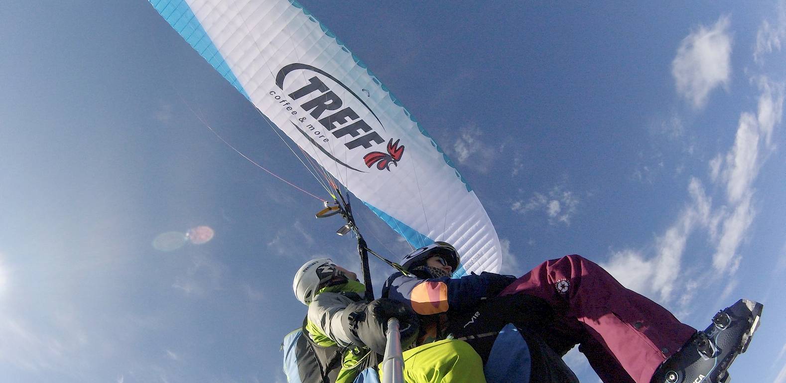 Due persone in parapendio biposto sotto un cielo blu con una vela bianca.