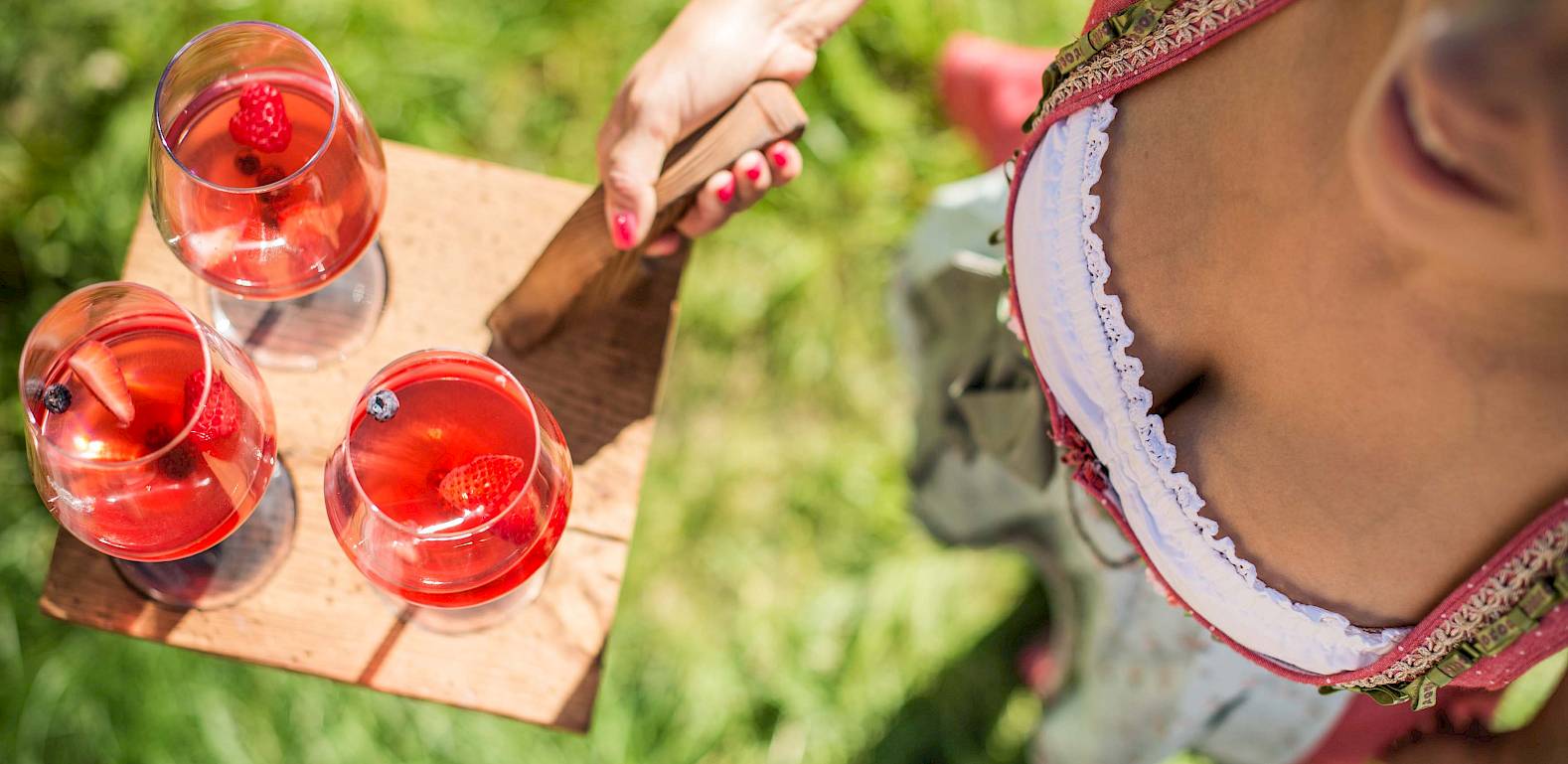 Frau im Dirndl hält Tablett mit drei Gläsern roter Fruchtbowle im Grünen.