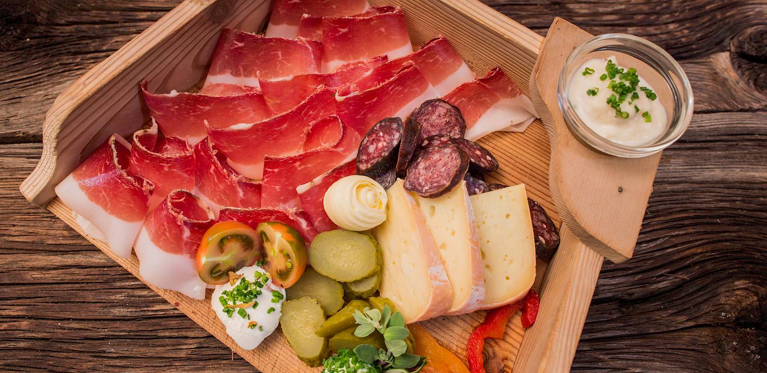 Holzplatte mit Schinken, Salami, Käse, Tomaten, Gurken und Dip. Rustikaler Hintergrund aus Holz.