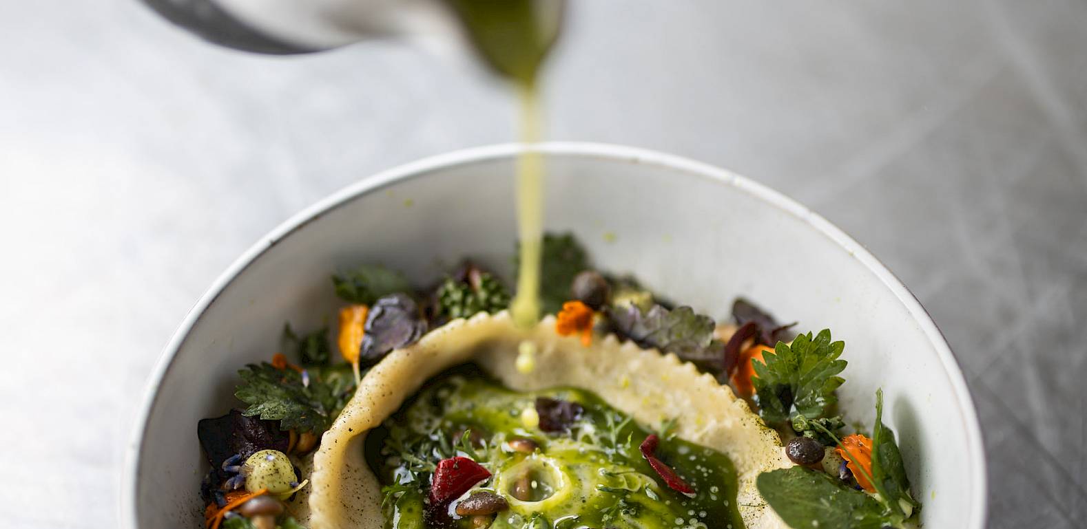 A hand pours green sauce over creatively presented appetizer with vegetables and edible flowers.