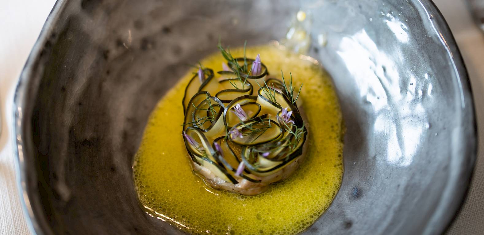A gourmet dish in a gray bowl is being topped with yellow sauce poured from a jug.