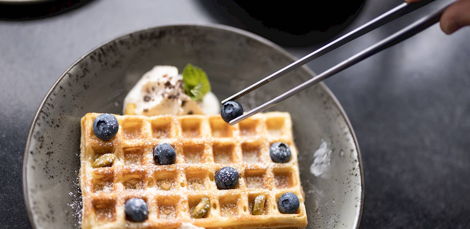 Waffel mit Heidelbeeren und Sahne auf einem Teller. Eine Person fügt Heidelbeeren mit einer Zange hinzu.