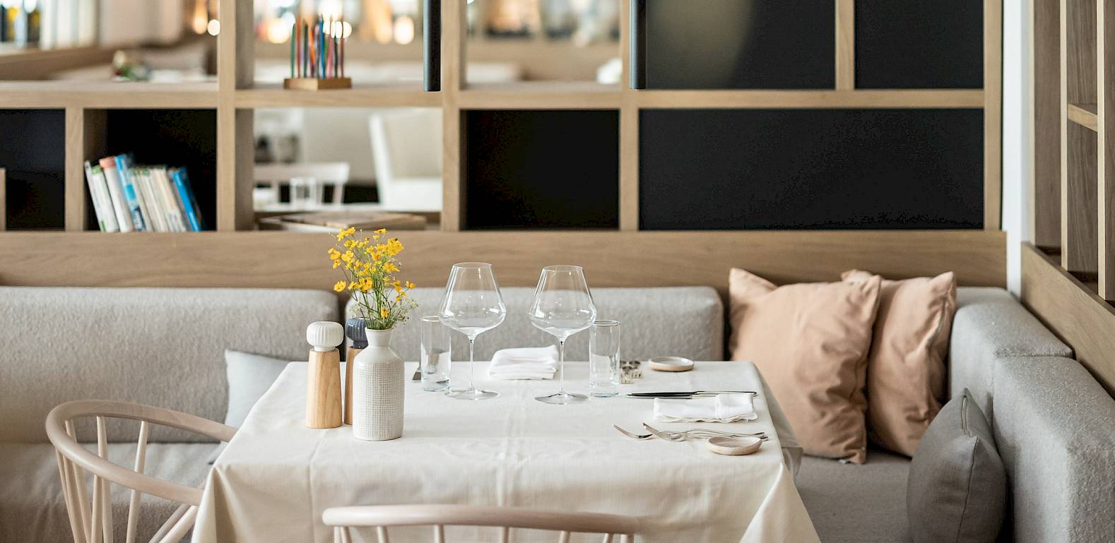 Gemütlich gedeckter Tisch in modernem Restaurant mit Gläsern, Kissen und Blick auf die Bar im Hintergrund.